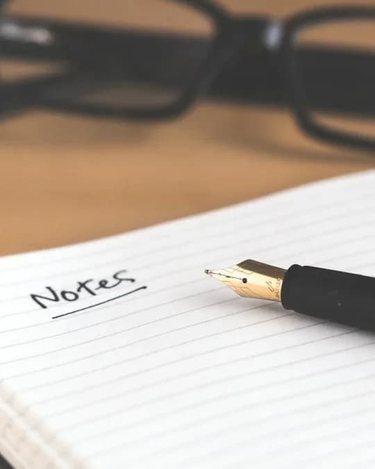 Caderno aberto com a palavra “Notes”, uma caneta-tinteiro e óculos sobre a mesa