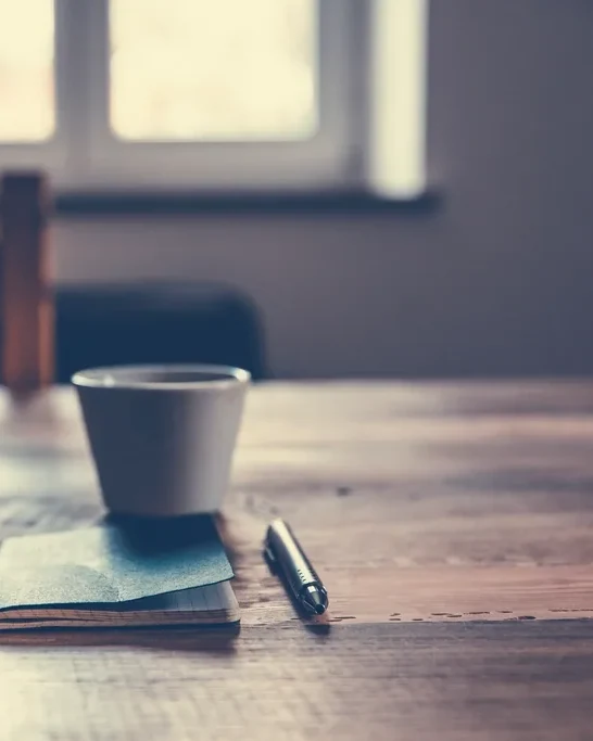 Copo de café sobre um bloco de notas e uma caneta, numa mesa de madeira perto da janela